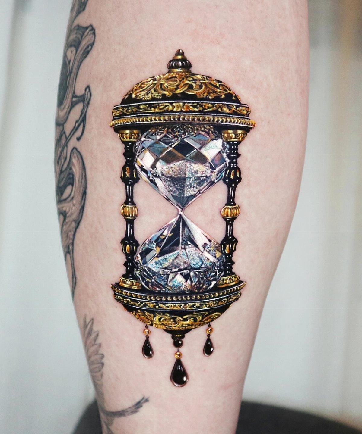 Ornate gold and black hourglass tattoo with diamond crystals on a person's leg