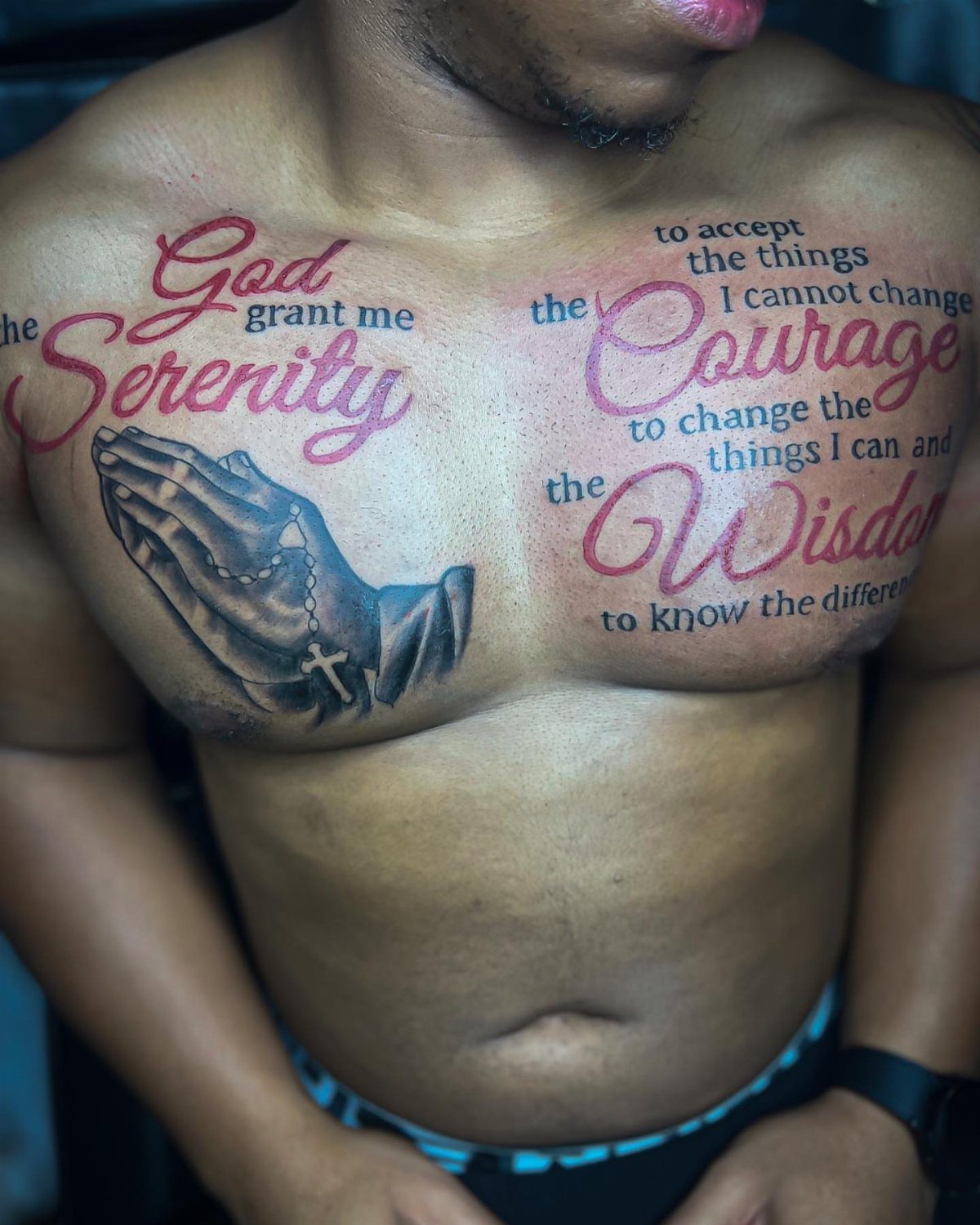 Chest tattoos of the Serenity Prayer in red script and praying hands.