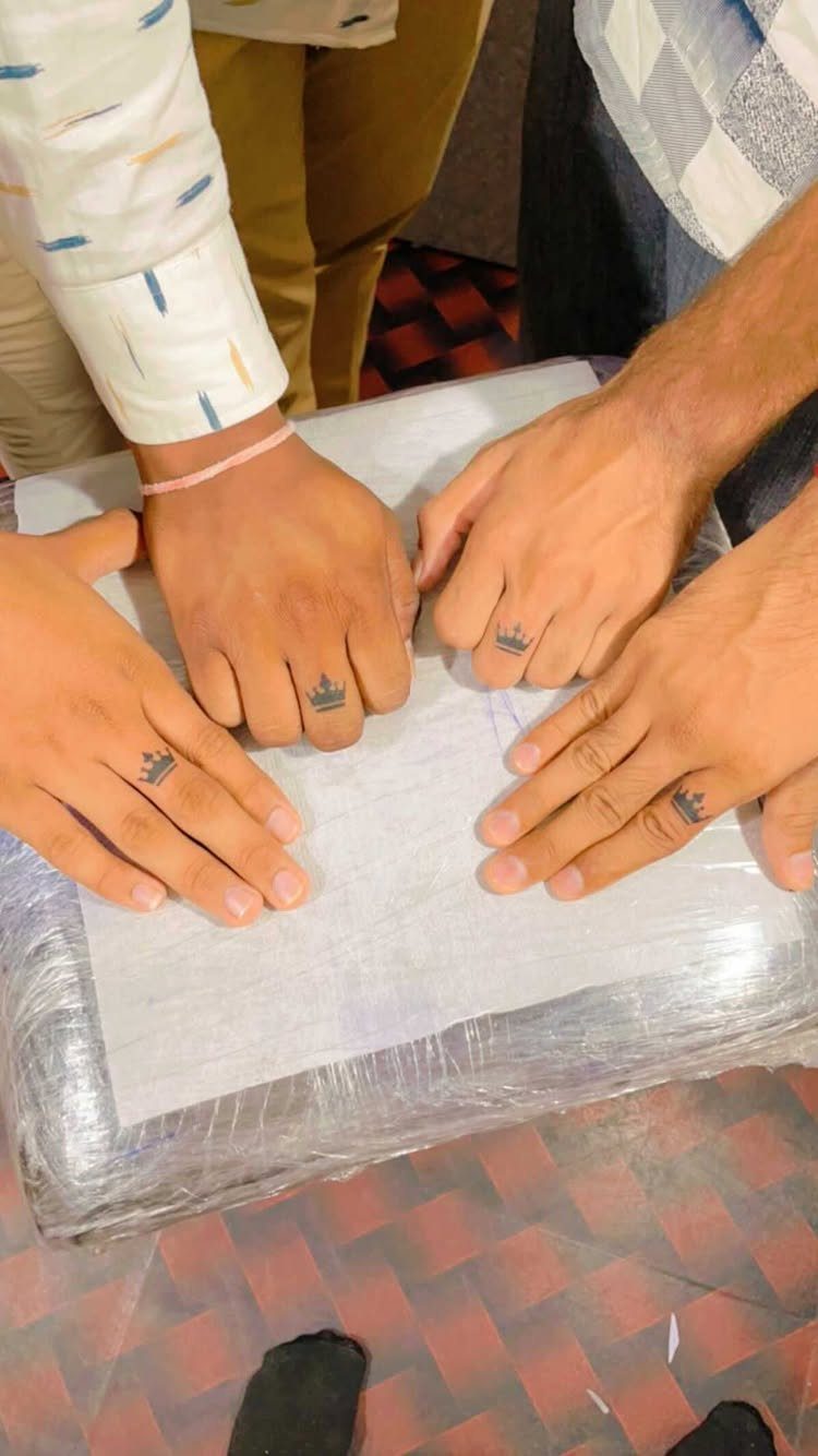 Four hands with small black crown tattoos on the ring fingers resting together.
