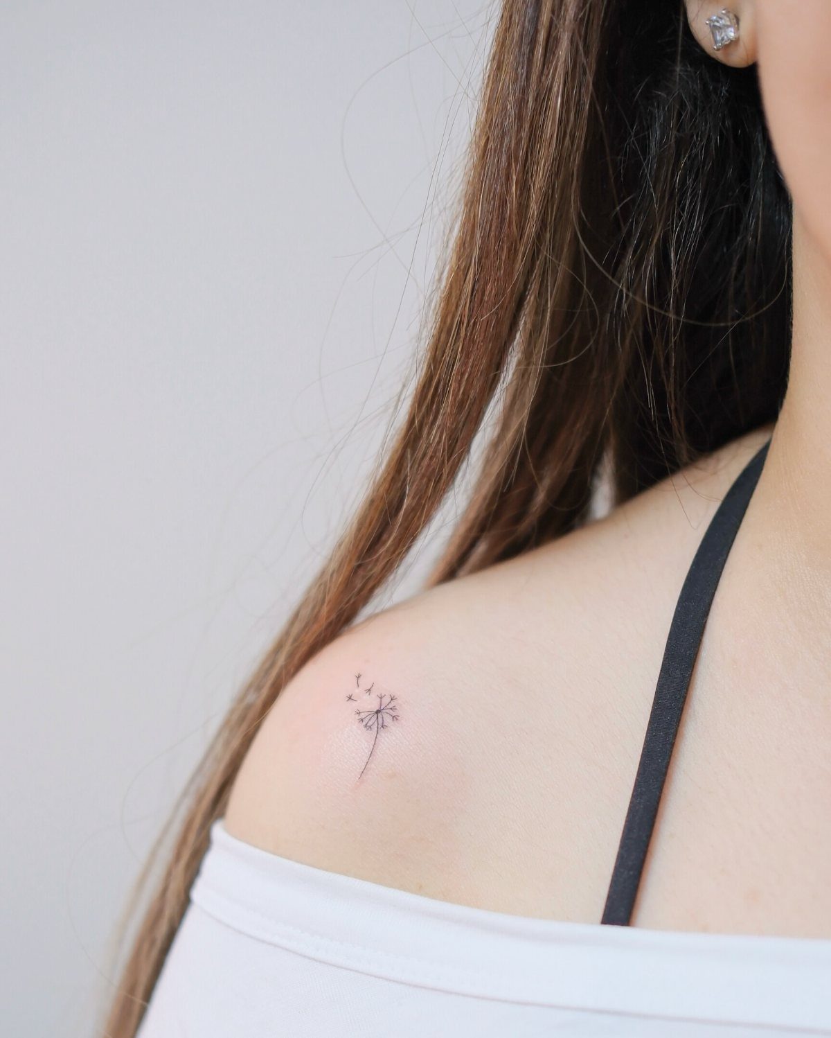 Small minimalist dandelion tattoo on a person's shoulder with floating seeds.