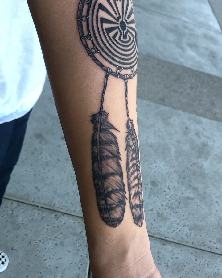 Dreamcatcher tattoo with two feathers on a person's forearm against concrete pavement.