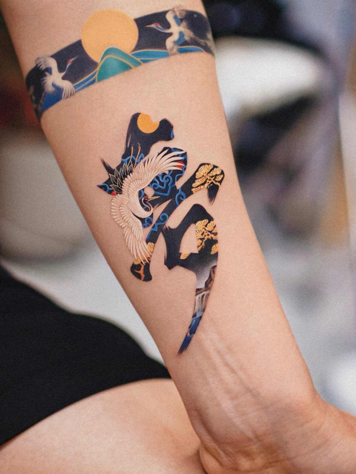 Forearm with colorful tattoos featuring a crane, sun, and Japanese-style landscapes