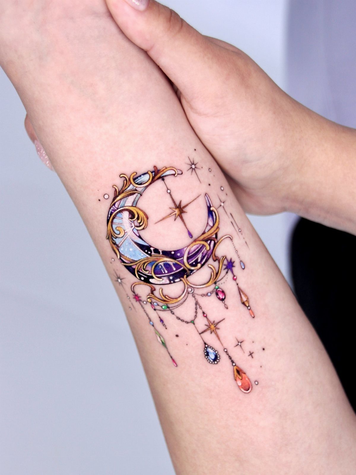 Crescent moon tattoo with gold filigree and colorful jewels on a forearm