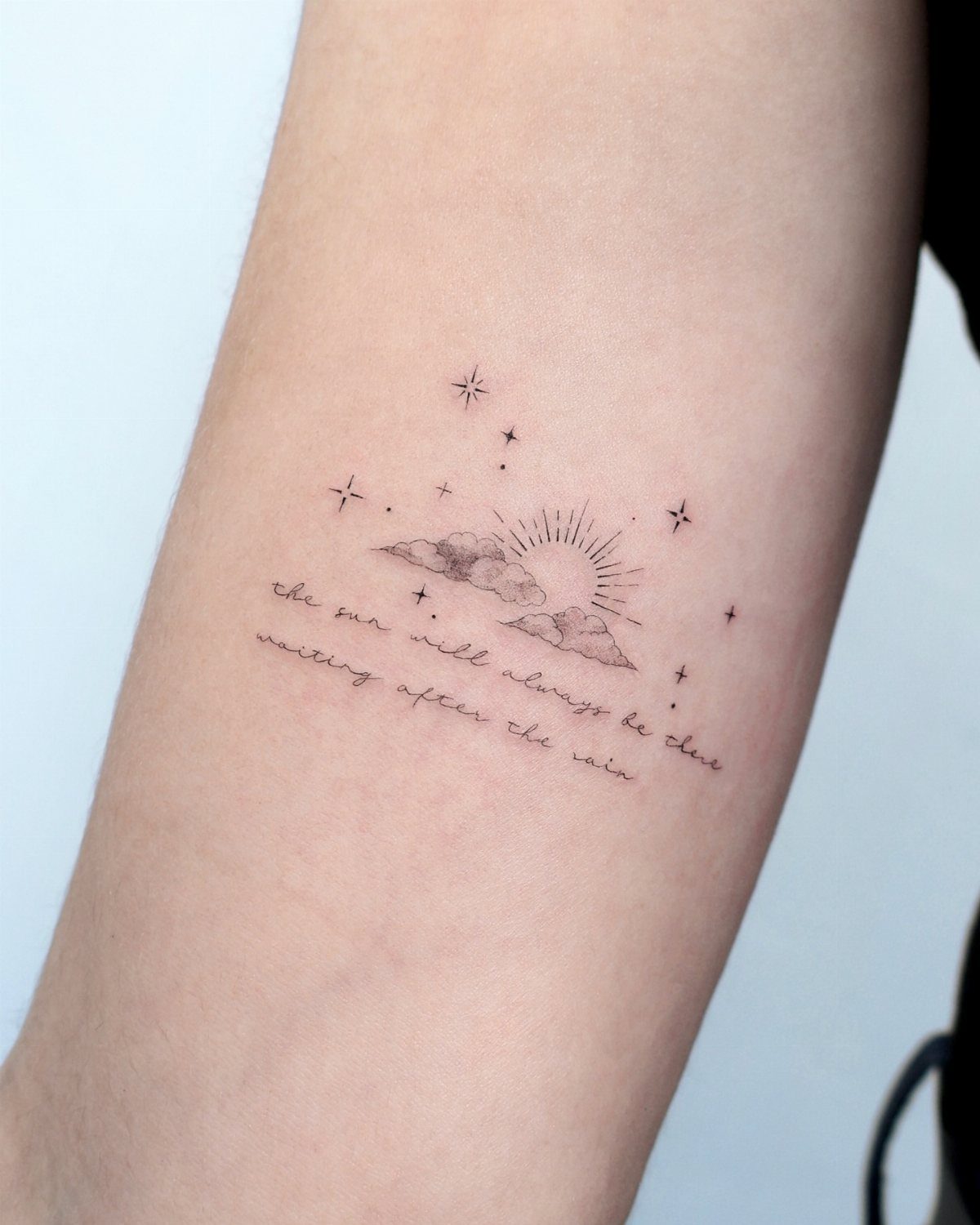 Fine-line tattoo of clouds, a sun, and stars with a cursive quote.