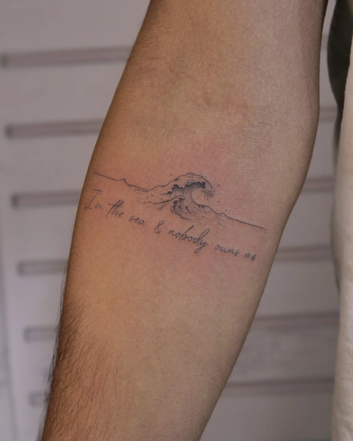 Forearm tattoo of a crashing ocean wave with a cursive quote underneath.