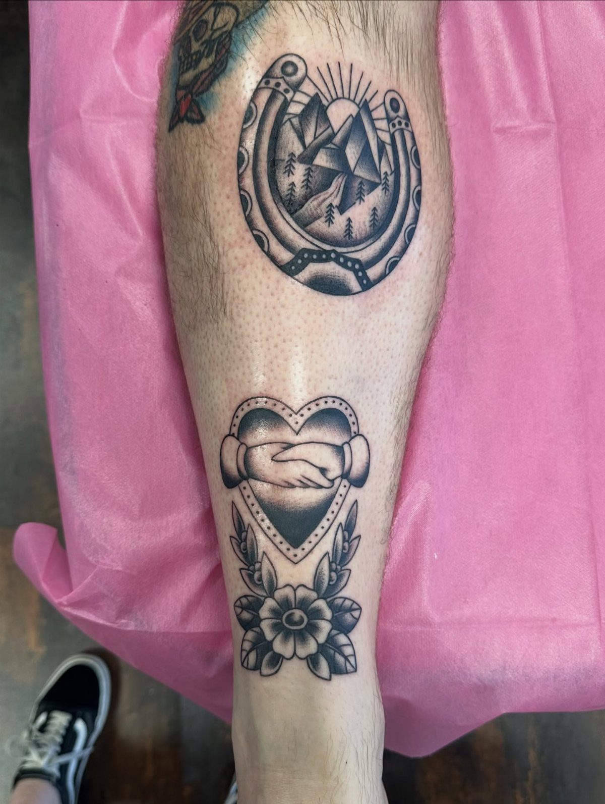 Traditional American tattoos of a horseshoe and shaking hands on a lower leg.