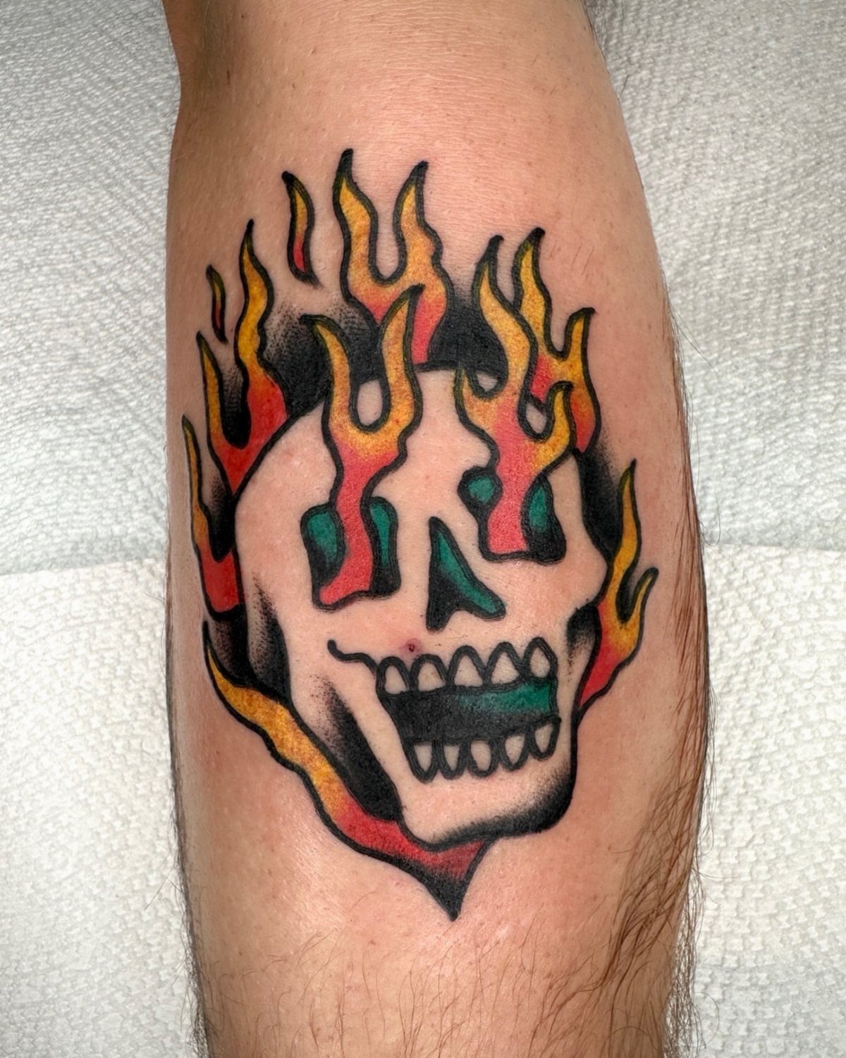 Flaming skull tattoo with thick black outlines and bold colors on a leg.