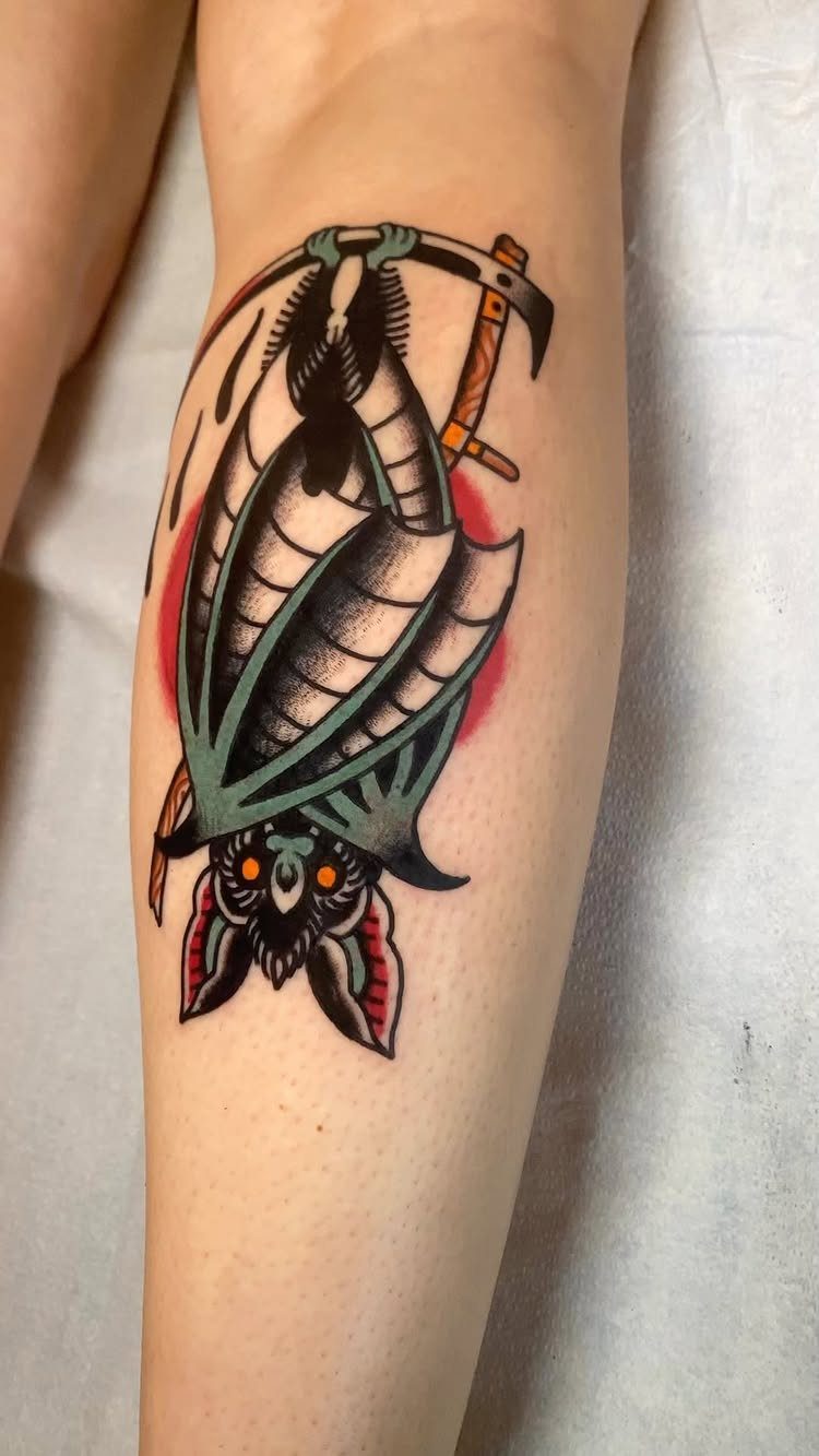 Upside-down bat tattoo with bold black outlines and red shading on a leg.