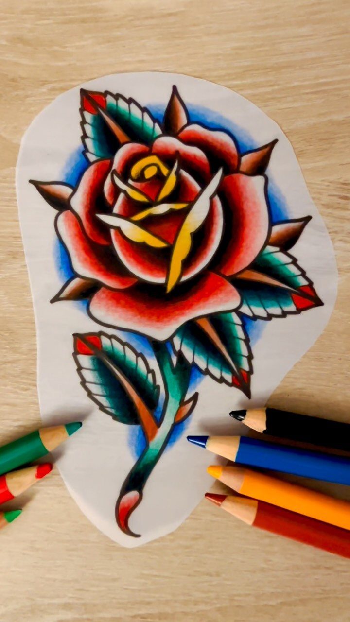Red rose tattoo design with bold outlines and colored pencils on wooden table