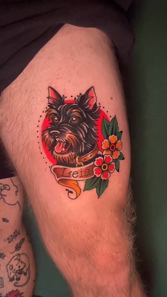 Traditional style tattoo of a black terrier dog with flowers and a banner.