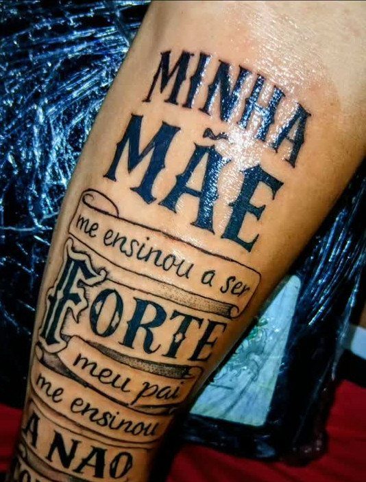 Tattoo on a forearm with bold traditional lettering and decorative parchment banners.