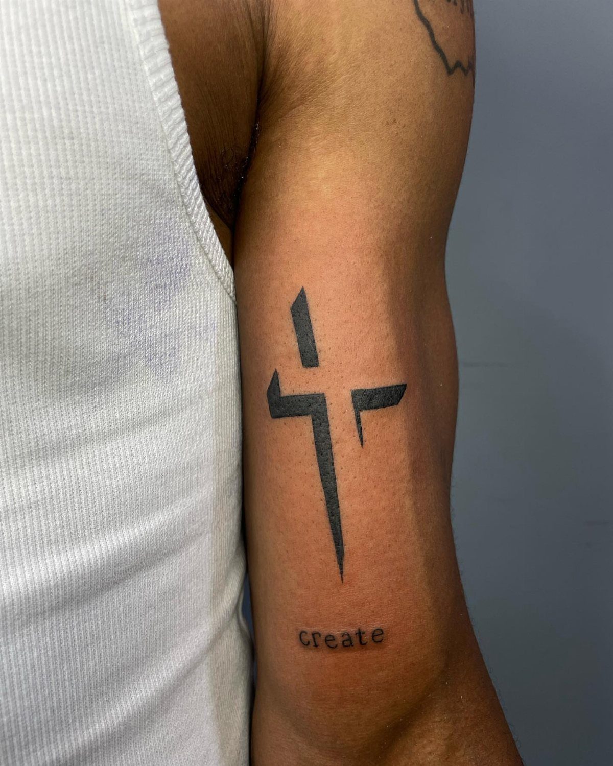 Stylized black tribal cross tattoo on a person's upper arm with text below.