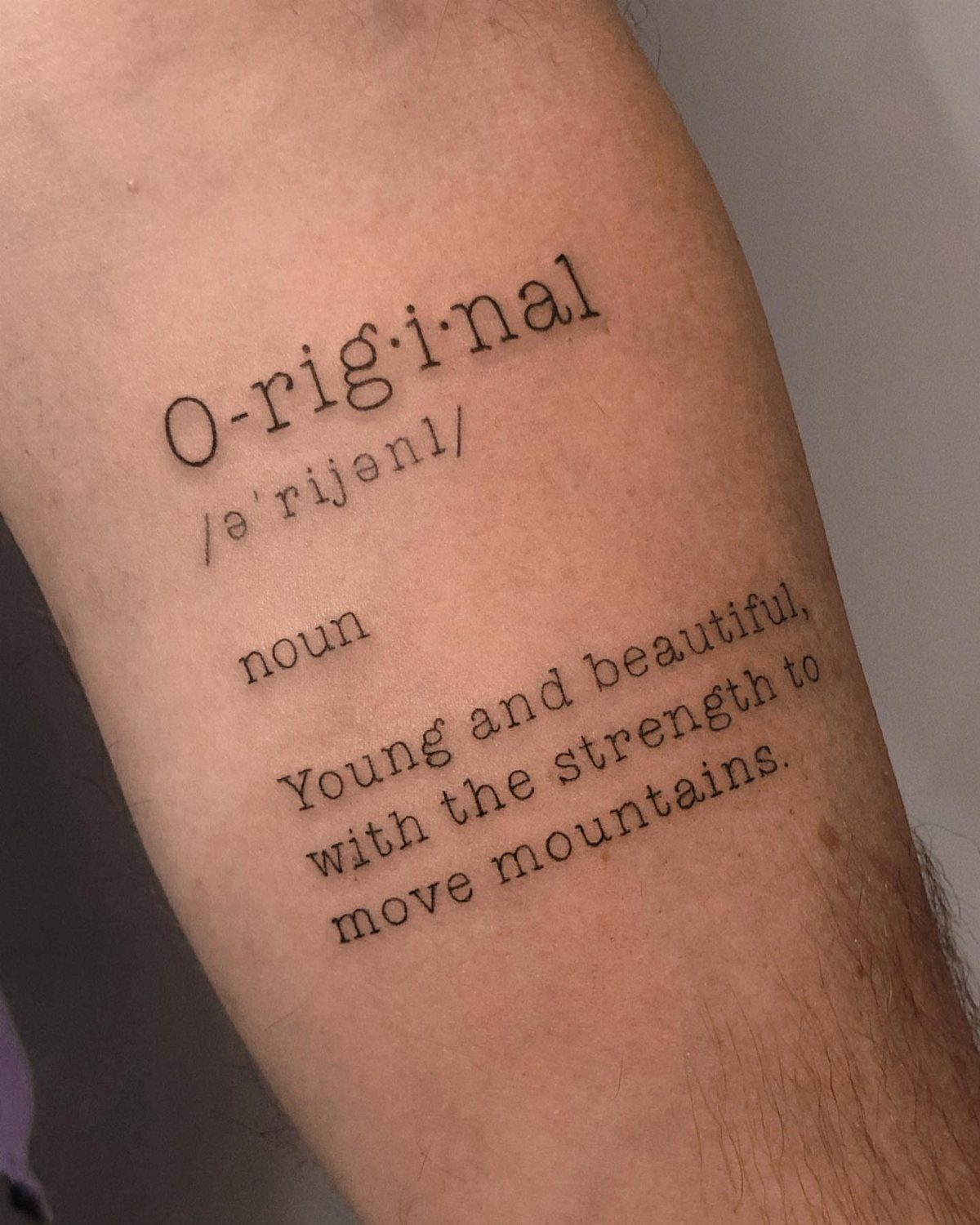 Typewriter font tattoo on a forearm defining the word original with a description.