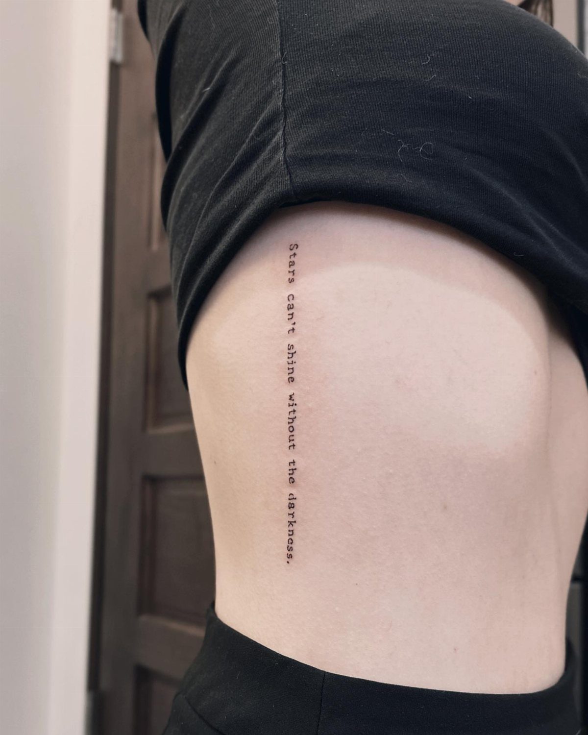 Typewriter font tattoo on a person's side reading Stars can't shine without the darkness.