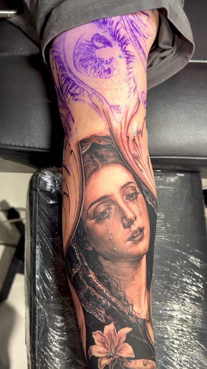 Crying Virgin Mary tattoo on a forearm with a lily flower below