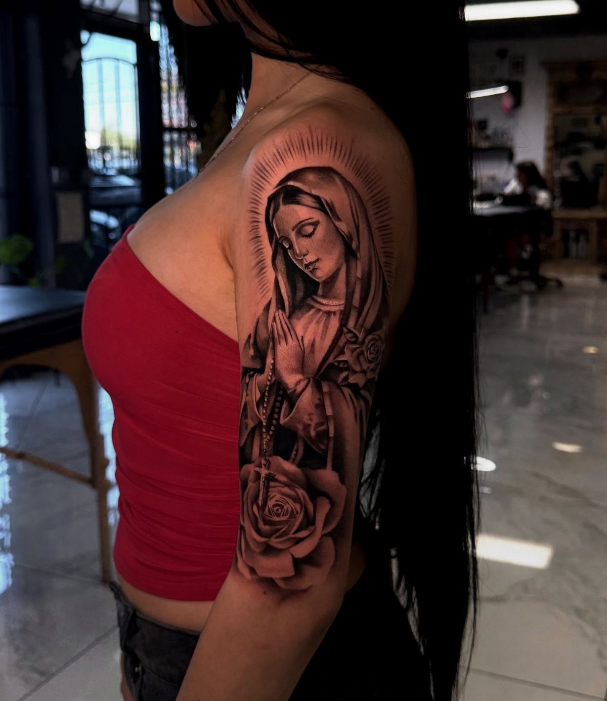 Virgin Mary tattoo on upper arm featuring a halo, rosary, and a rose.