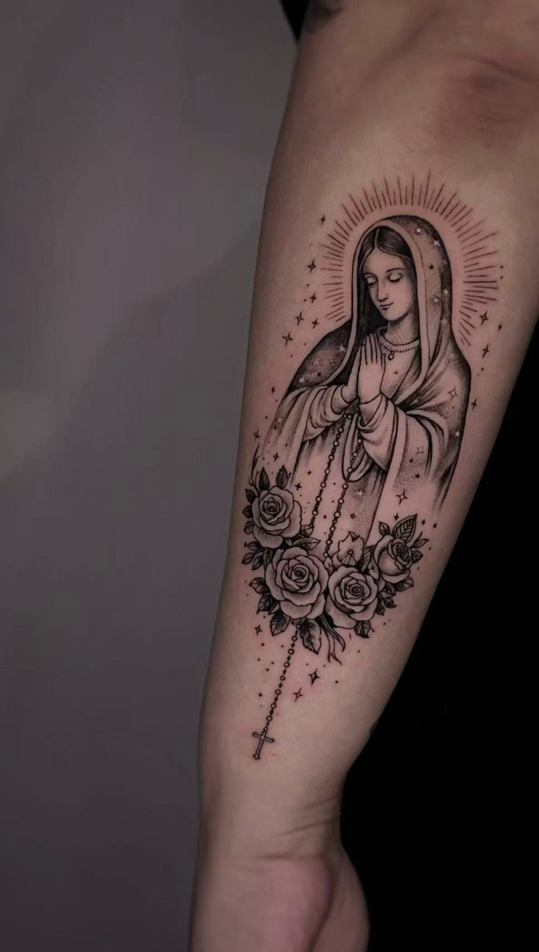 Virgin Mary tattoo on forearm featuring praying hands, roses, and a rosary.