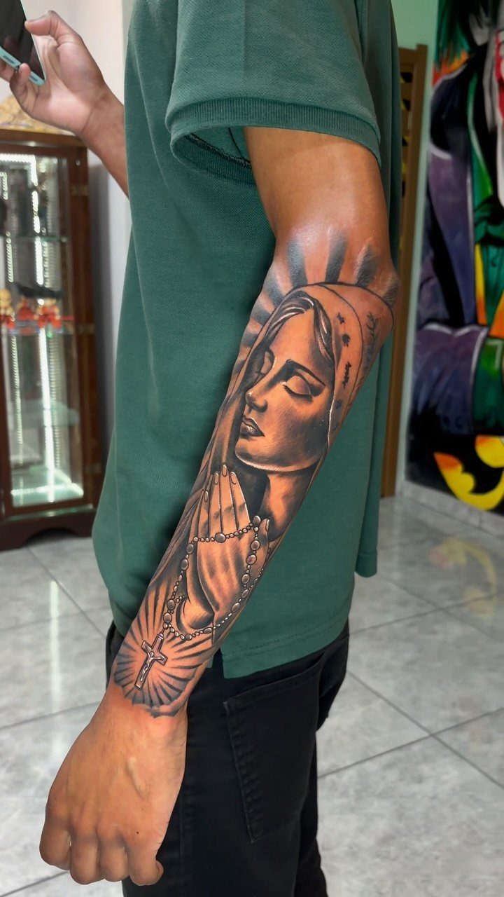 Virgin Mary tattoo on a forearm featuring praying hands, rosary, and radiating light rays.