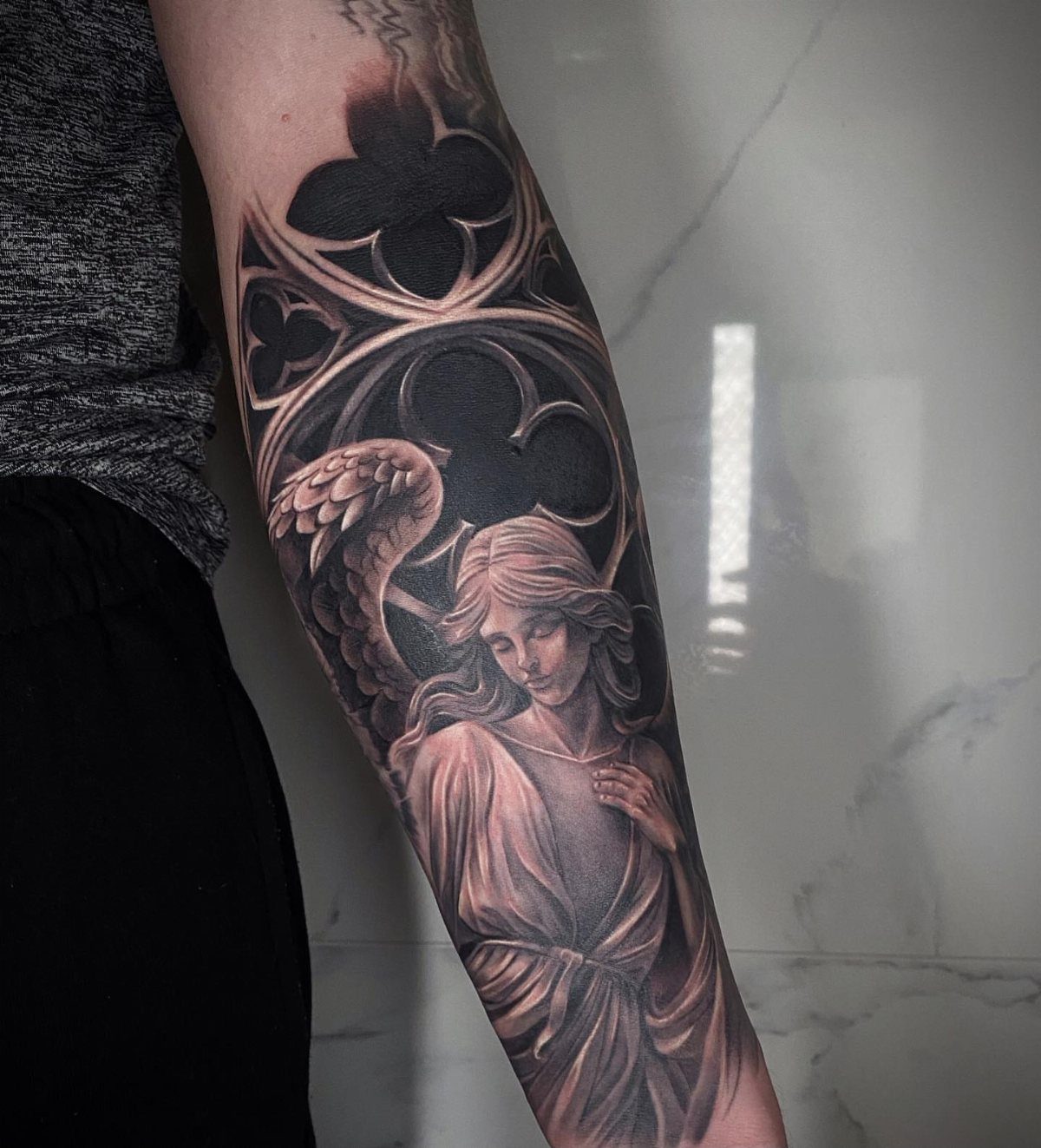 Detailed black and grey angel tattoo on a forearm with gothic architectural elements.
