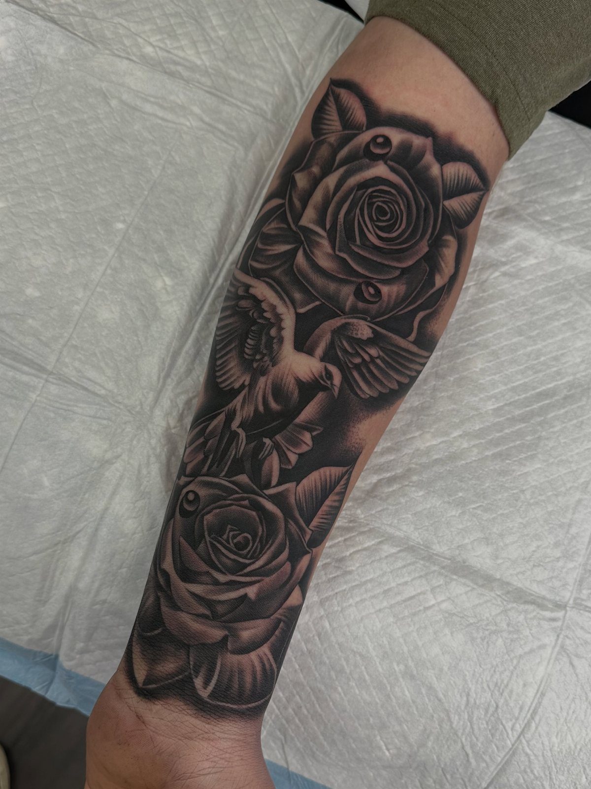 Dove flying between two roses in a black and grey forearm tattoo.