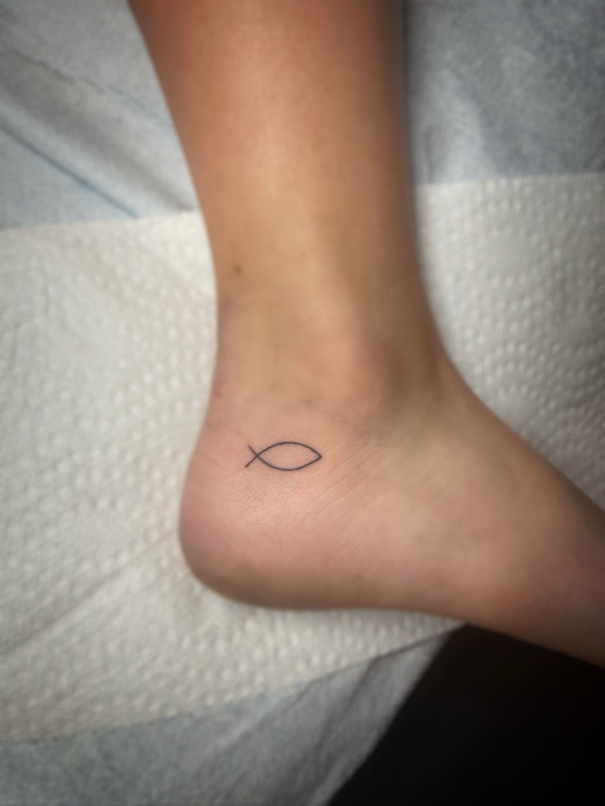 Small black Ichthys fish tattoo on the side of a person's ankle.