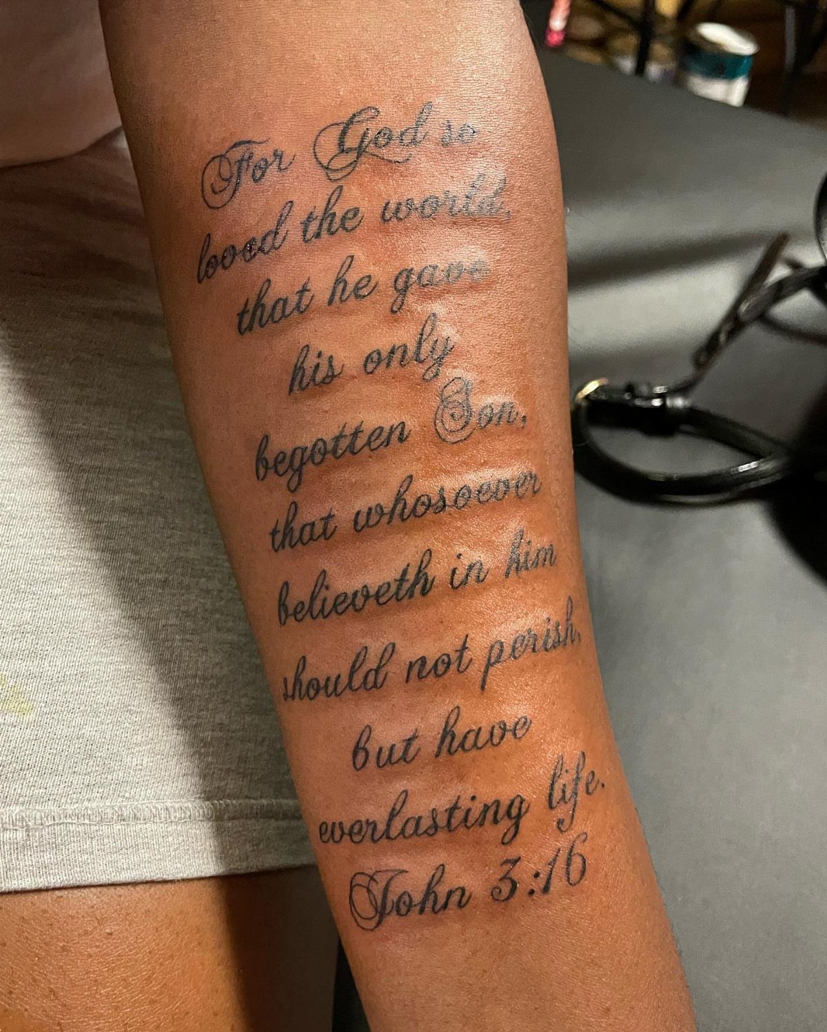 Forearm tattoo featuring the full text of John 3:16 in elegant black cursive script.