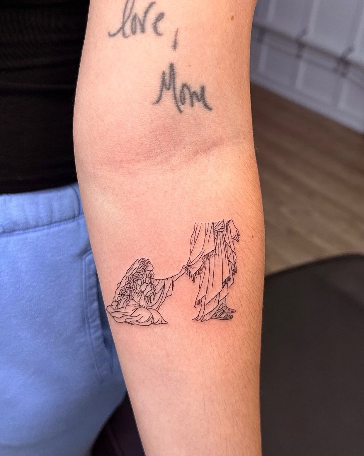 Fine-line tattoo of a woman kneeling before a robed figure on a forearm.