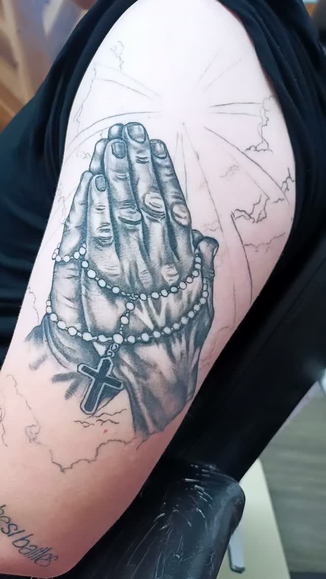 Praying hands tattoo with a rosary and cross on a person's upper arm.