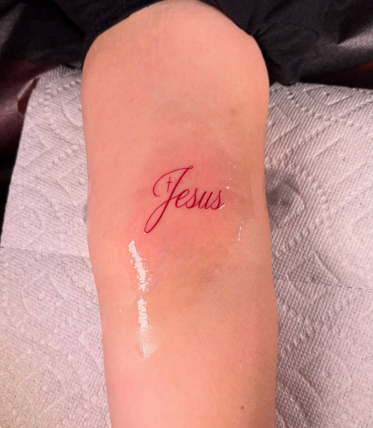 Red cursive Jesus tattoo with a small cross on a person's forearm.