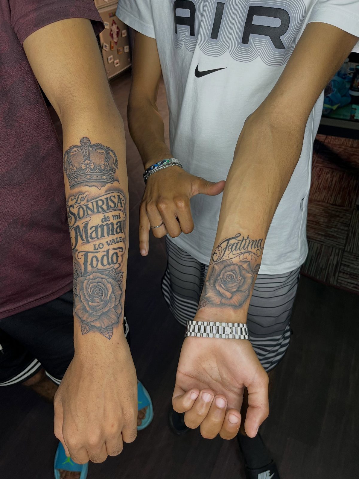 Two forearms displaying black ink tattoos with Spanish text and rose designs.