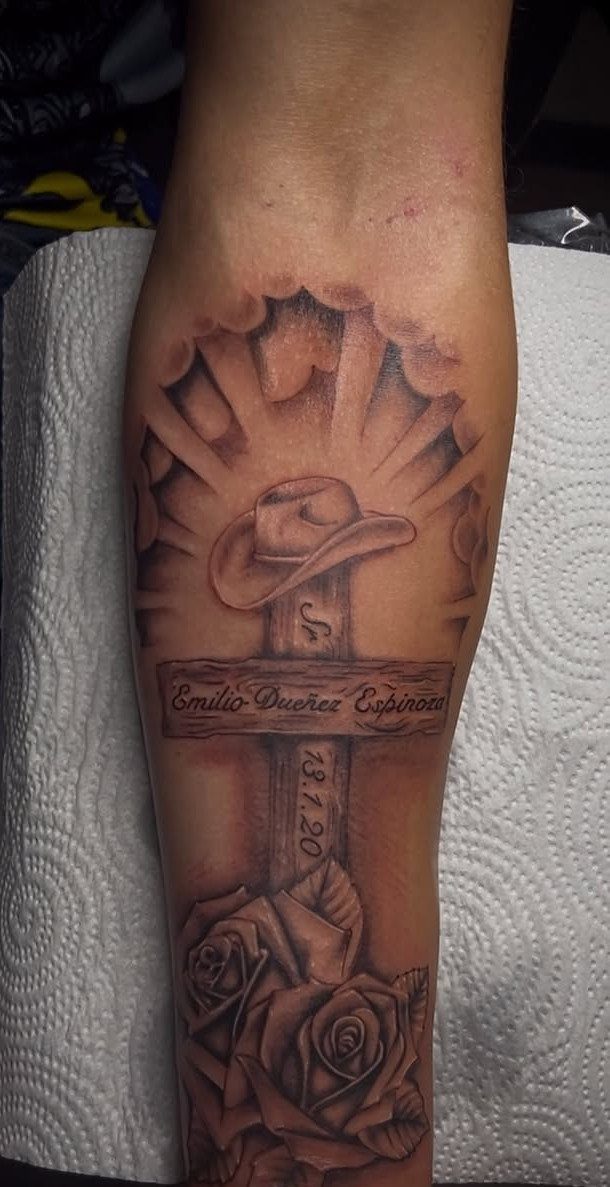 Memorial tattoo on forearm featuring a cross, cowboy hat, roses, and a name banner.