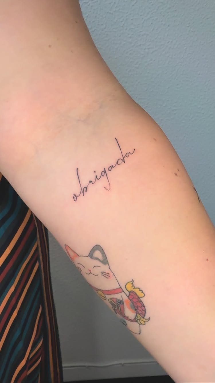 Cursive 'obrigada' tattoo and a lucky cat tattoo on a person's forearm.
